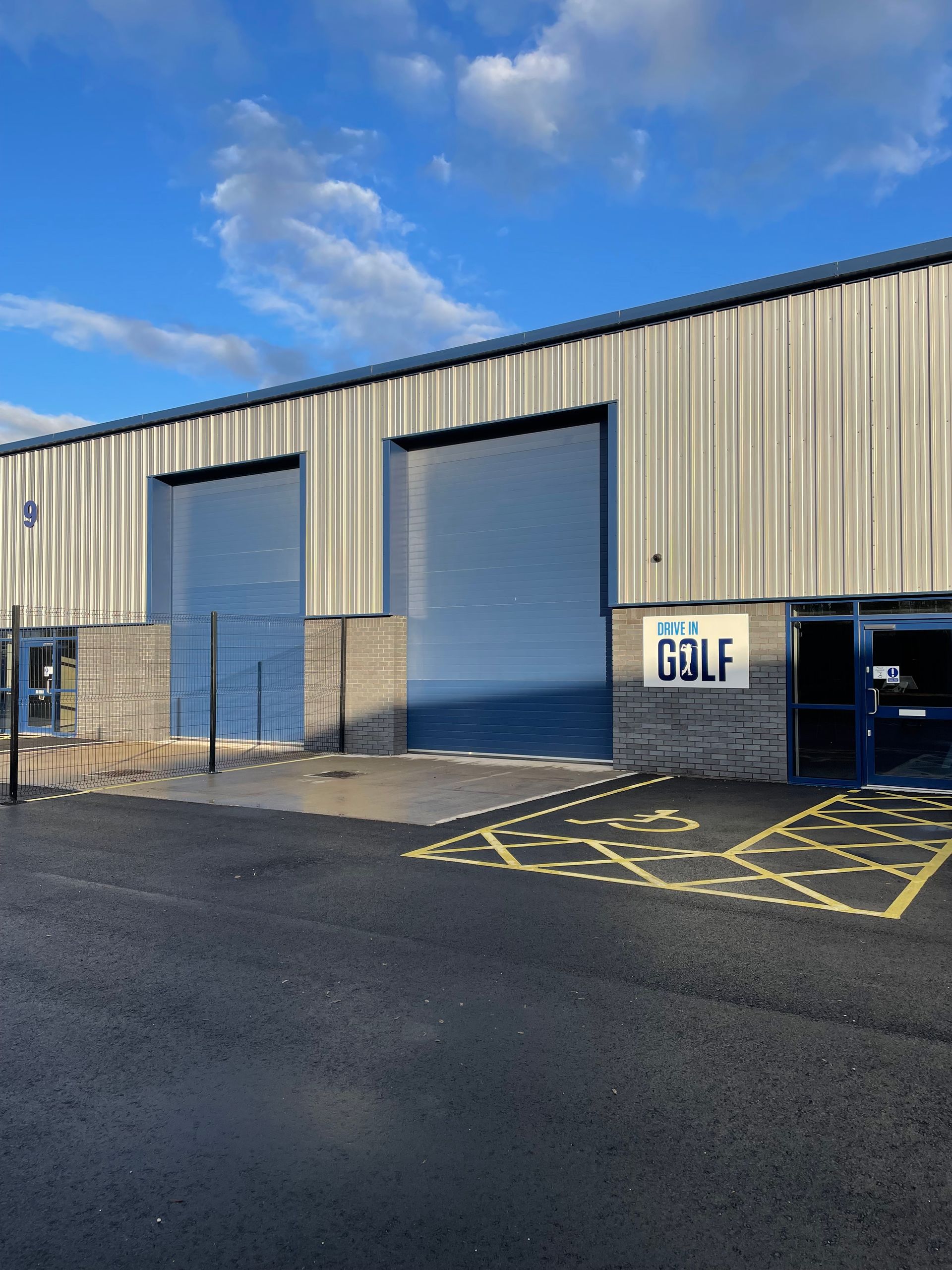 Industrial warehouse with blue roller doors, fenced loading area, and “GOLF” sign under a blue sky