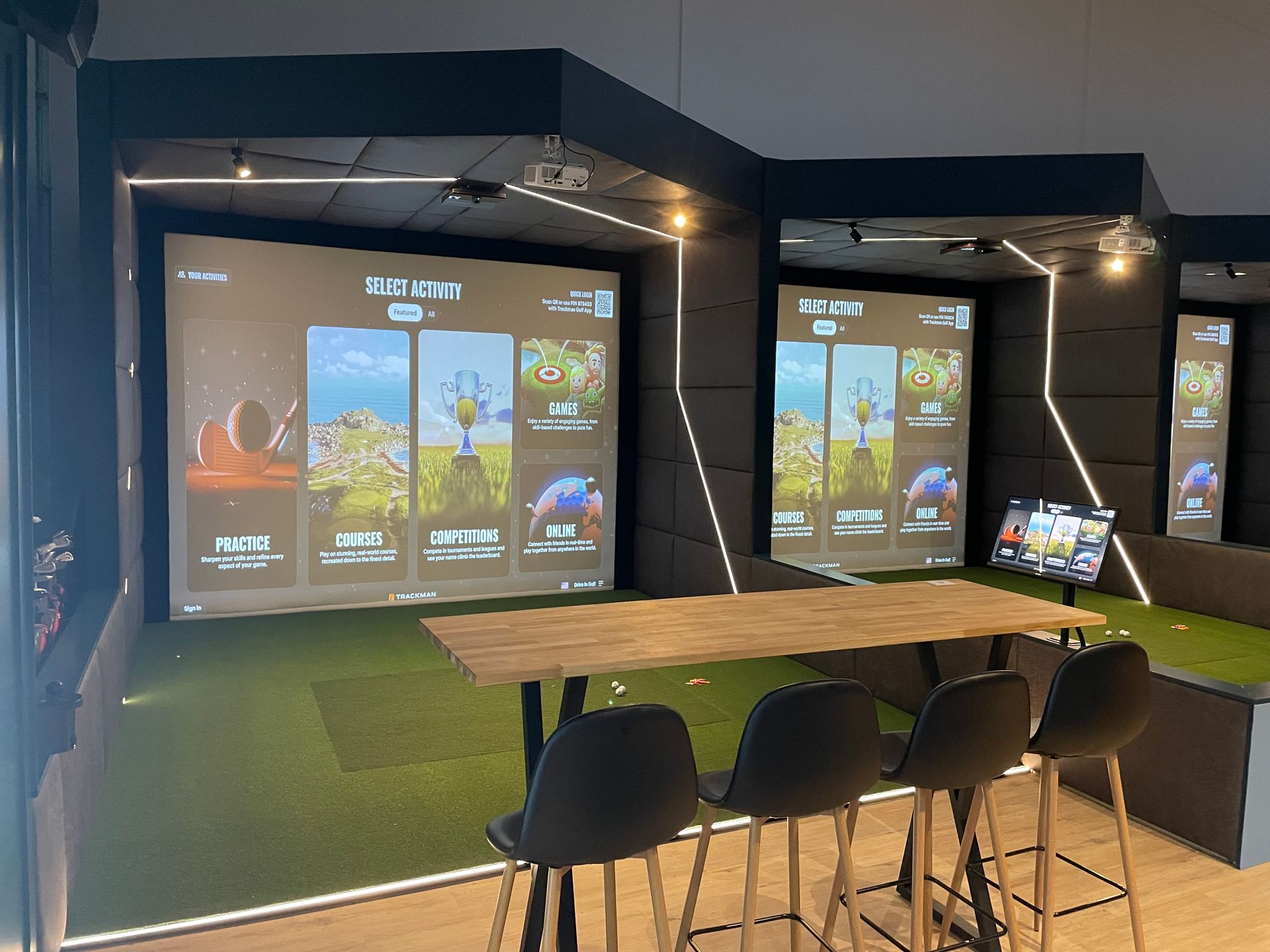 A cozy indoor gaming lounge with large wall screens, a wooden table, and black chairs under warm lighting.