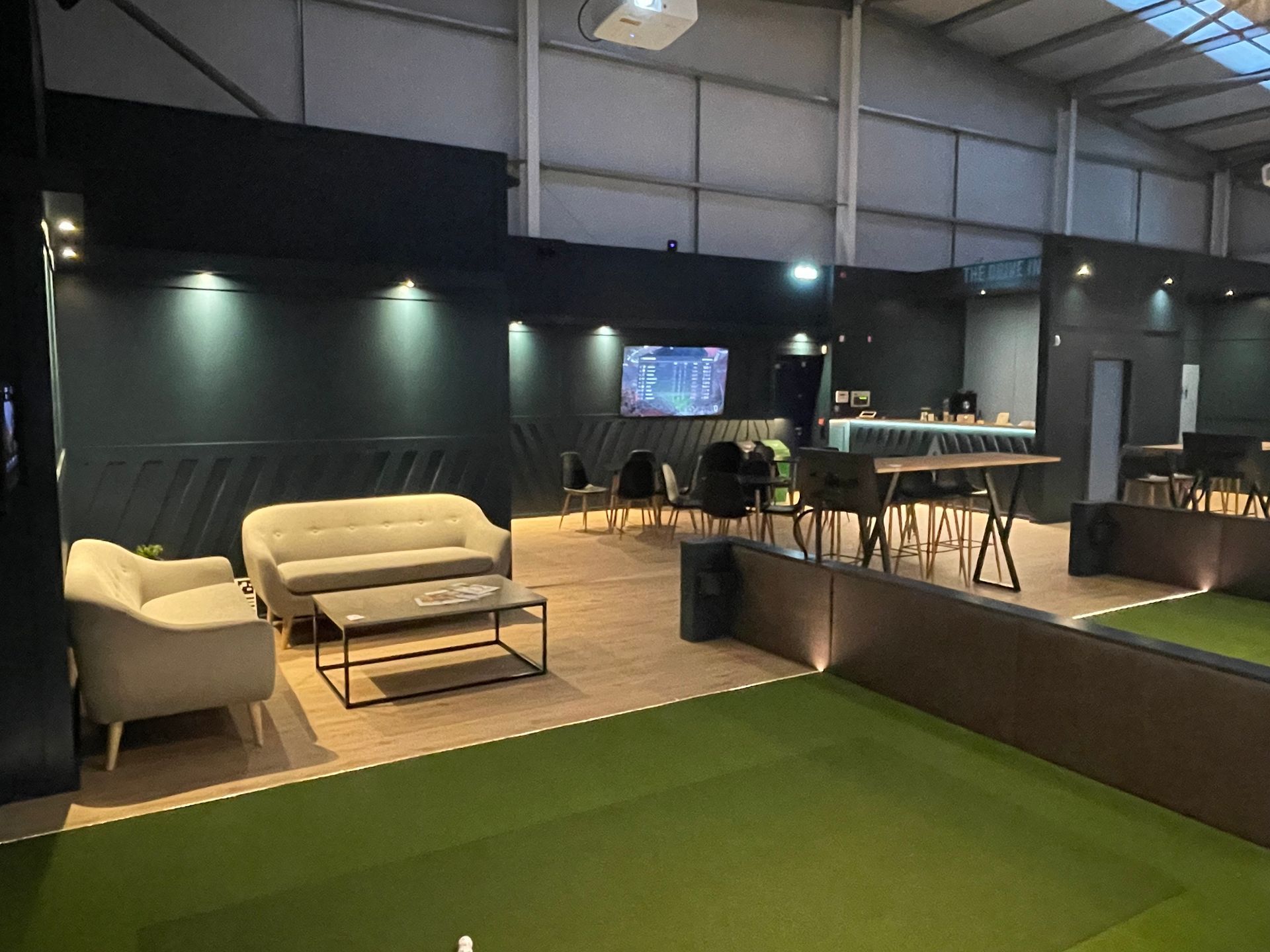 Modern lounge with sofas, bar tables, wall lights, and a green indoor turf area