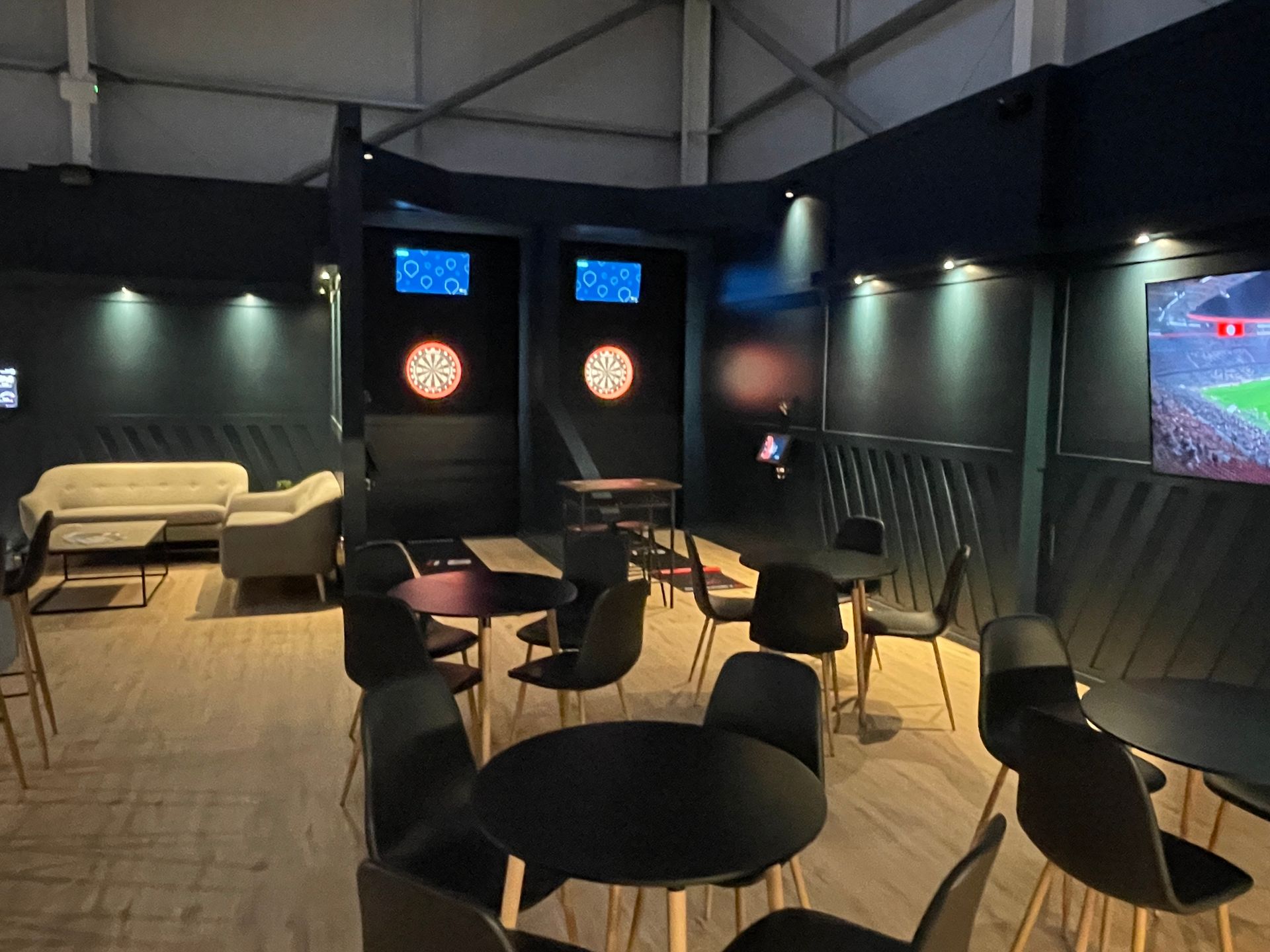 Empty lounge with round tables and chairs, wall-mounted TVs, and a beige sofa under dim lighting.