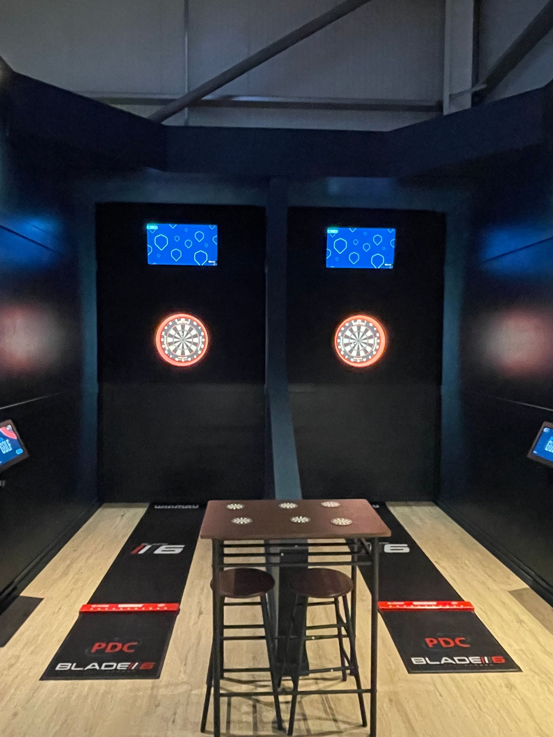 Indoor dart booth with two dartboards, overhead screens, and a narrow table with stools