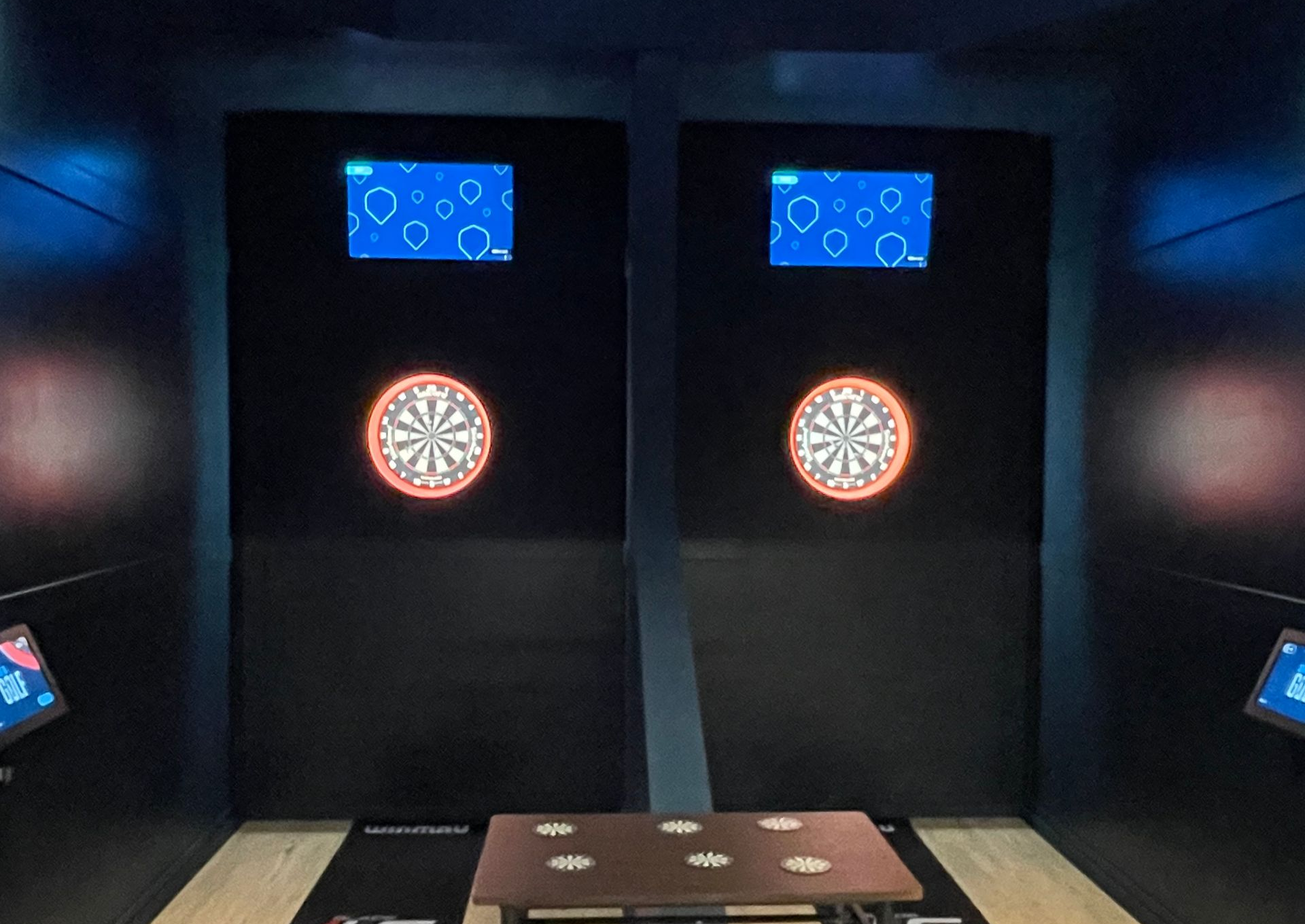 Indoor darts lane with two dartboards and blue score screens on a dark wall