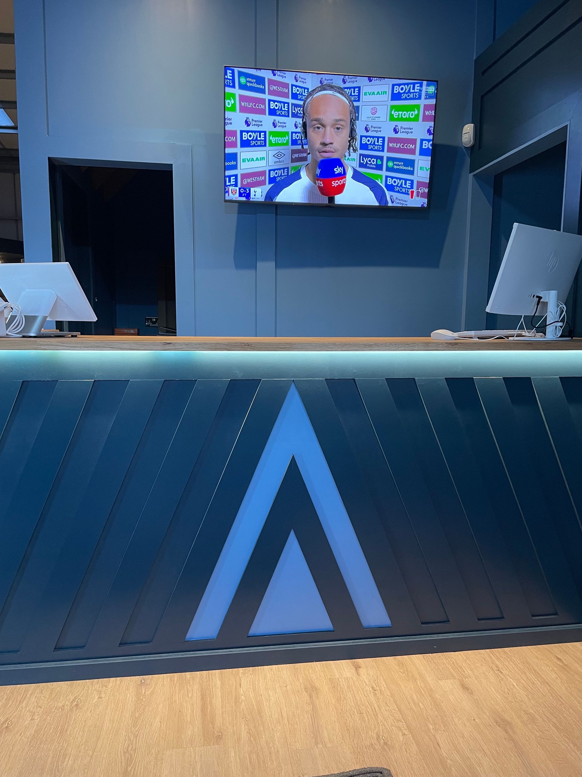 Modern reception desk with blue accent lighting and a wall-mounted TV showing a man speaking in a studio.