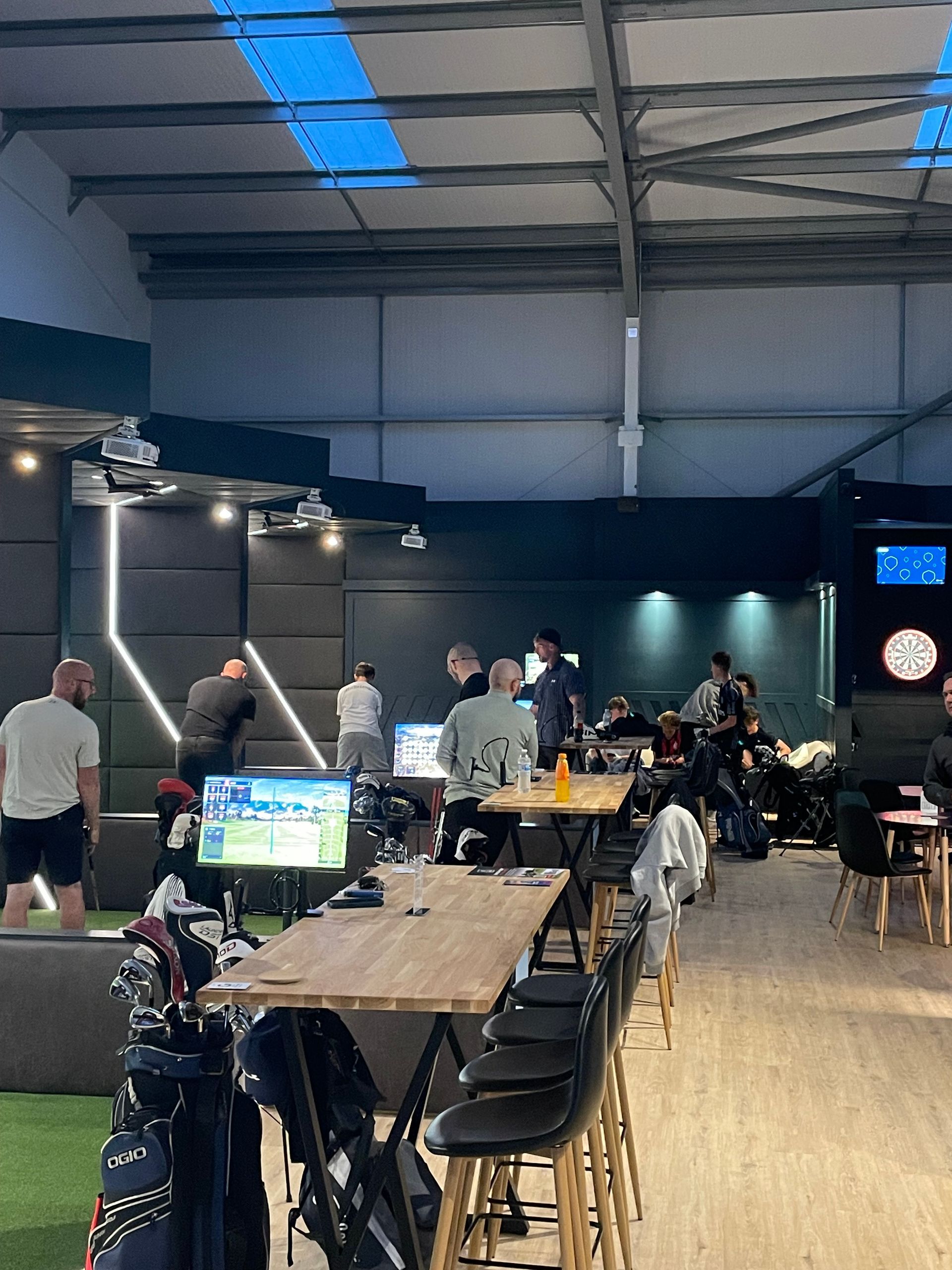 Indoor hall with tables, chairs, a dartboard, and people gathered near the back wall