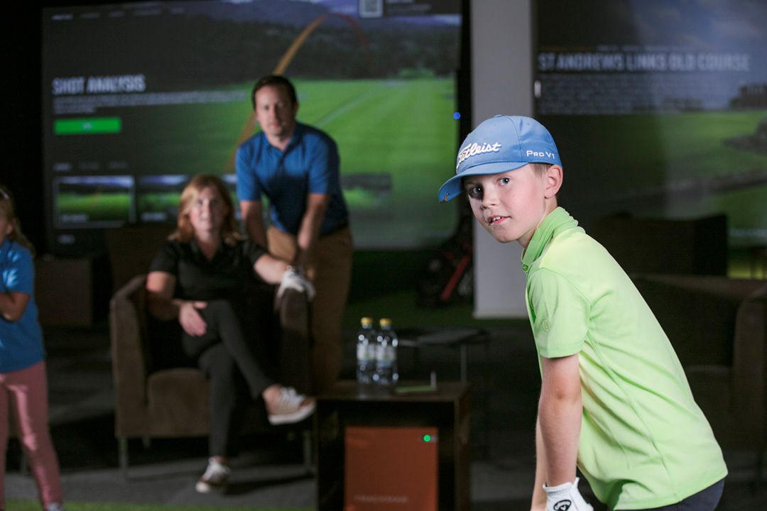 Boy in green shirt and blue cap at golf simulator; parents watch in background.
