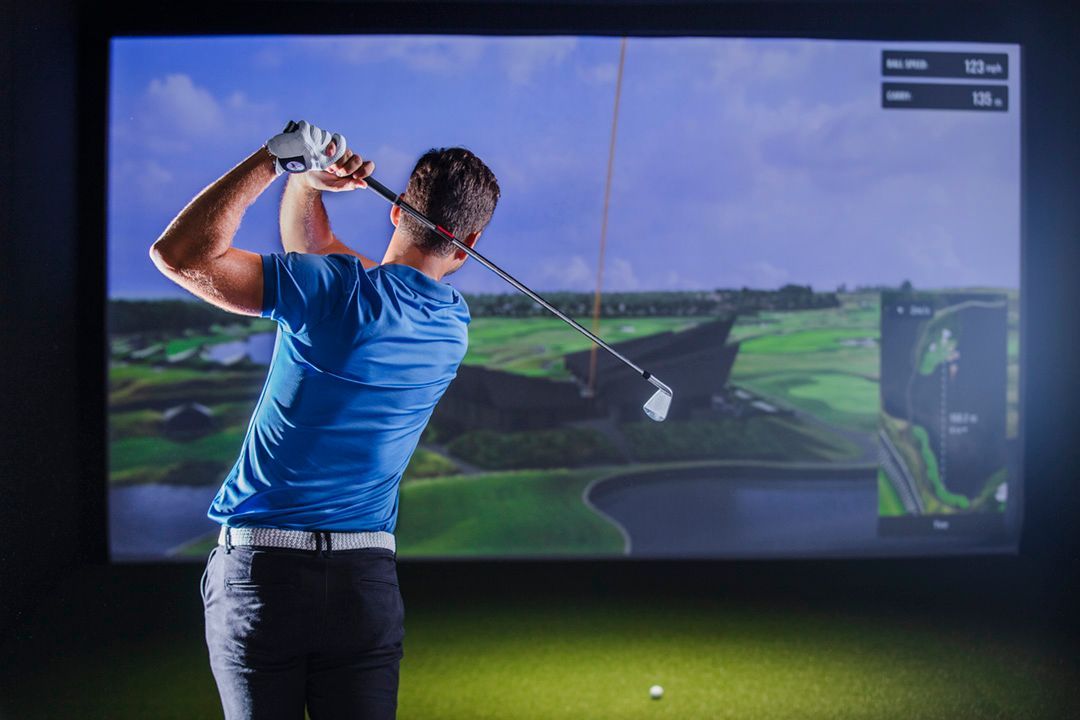 Golfer in a blue shirt swings in a golf simulator; screen shows a course.