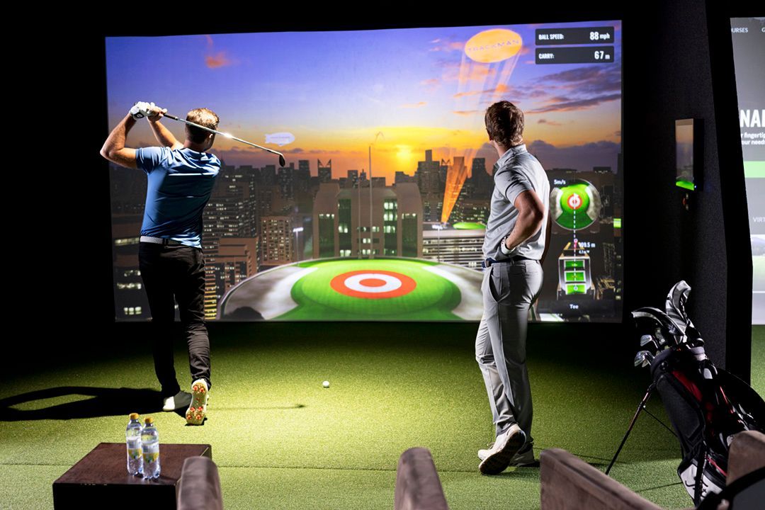 Two men in a golf simulator: one swings, the other watches, a cityscape on the screen behind them.