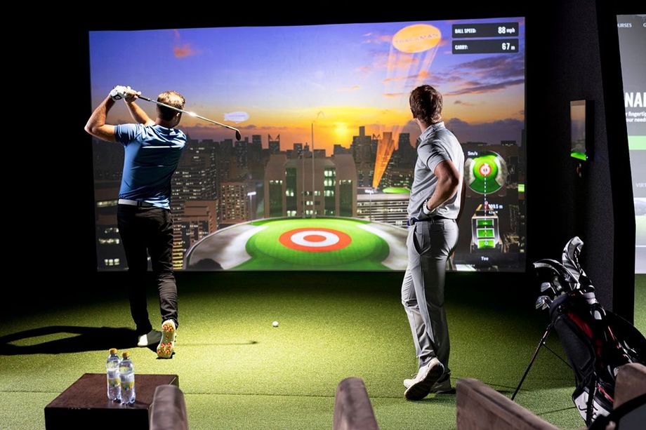 Man swings golf club indoors, facing projected course. Another man watches.