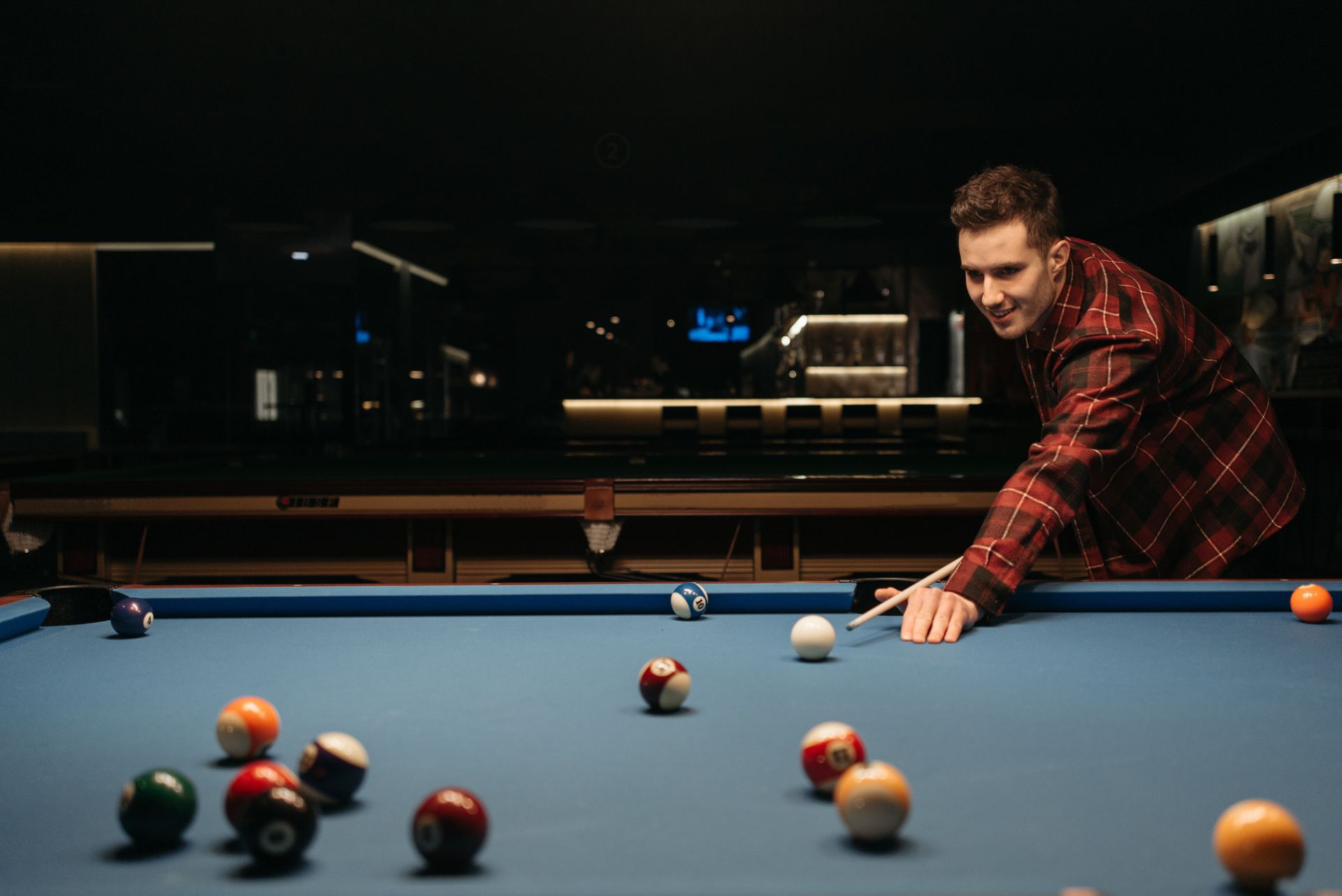 Man aiming a cue stick at a billiards ball on a blue pool table in a dimly lit room