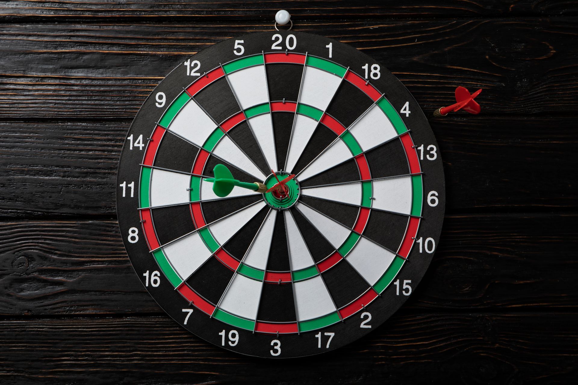 Dartboard with two darts: one in the center, and one near the top right. On a dark wooden background.