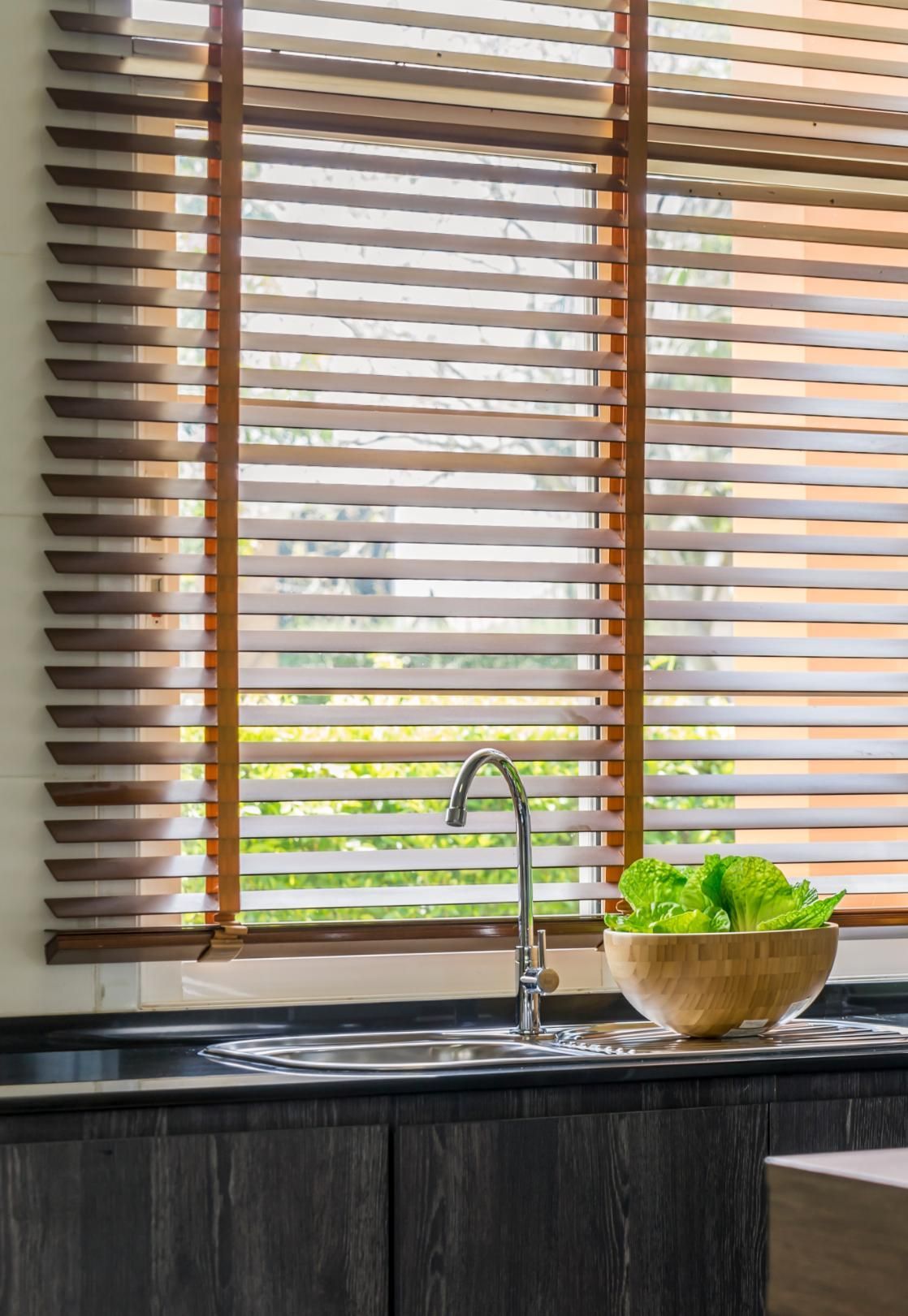 Kitchen sink by window with wooden blinds, chrome faucet, and bowl of green lettuce.