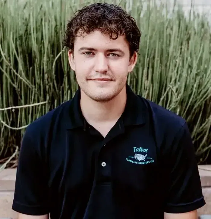 Young man in a black polo shirt seated in front of tall green plants, facing the camera.