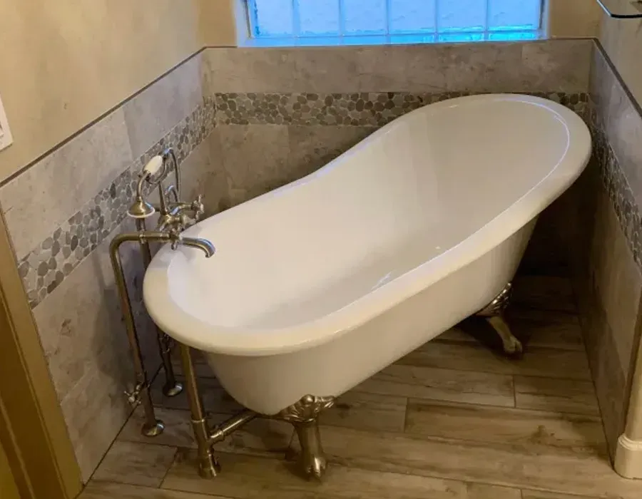 Freestanding white clawfoot bathtub in a small tiled bathroom with a window blind.