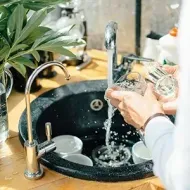 Hands washing dishes in a kitchen sink with running water and soap bubbles.