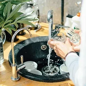 Hands washing dishes in a kitchen sink with running water and soap bubbles.