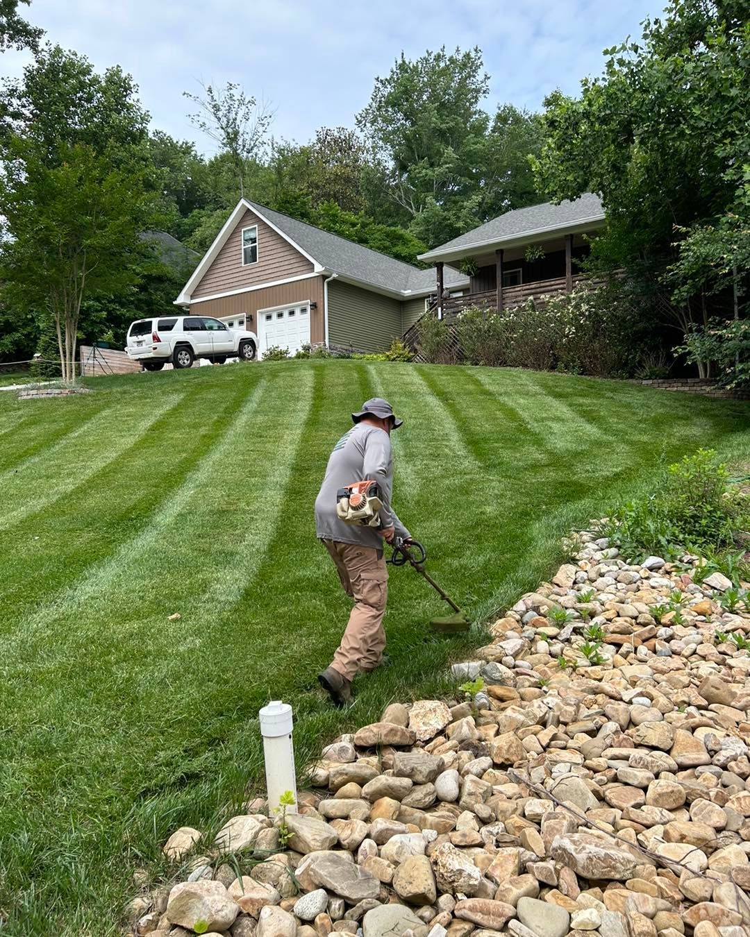 Person using a weed eater to trim grass  on a sloped lawn with striped patterns in front of a house.