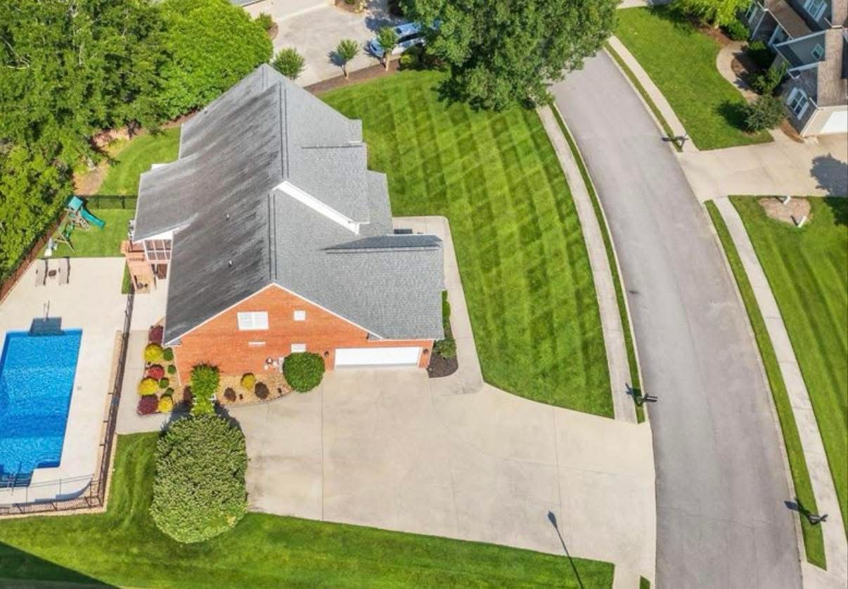 Aerial view of a suburban house with a blue pool, driveway, and curved street lined with trees.
