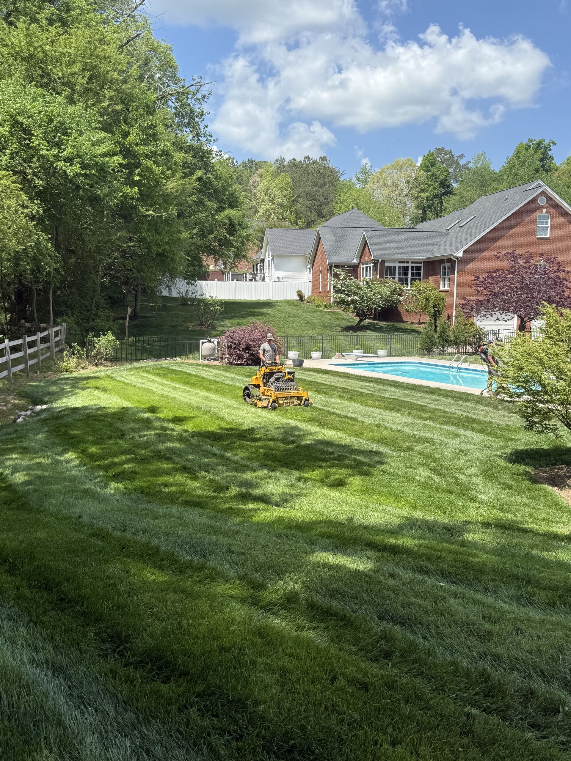 Backyard with green lawn, flower bed, trees, and a house beside a blue swimming pool under a partly cloudy sky