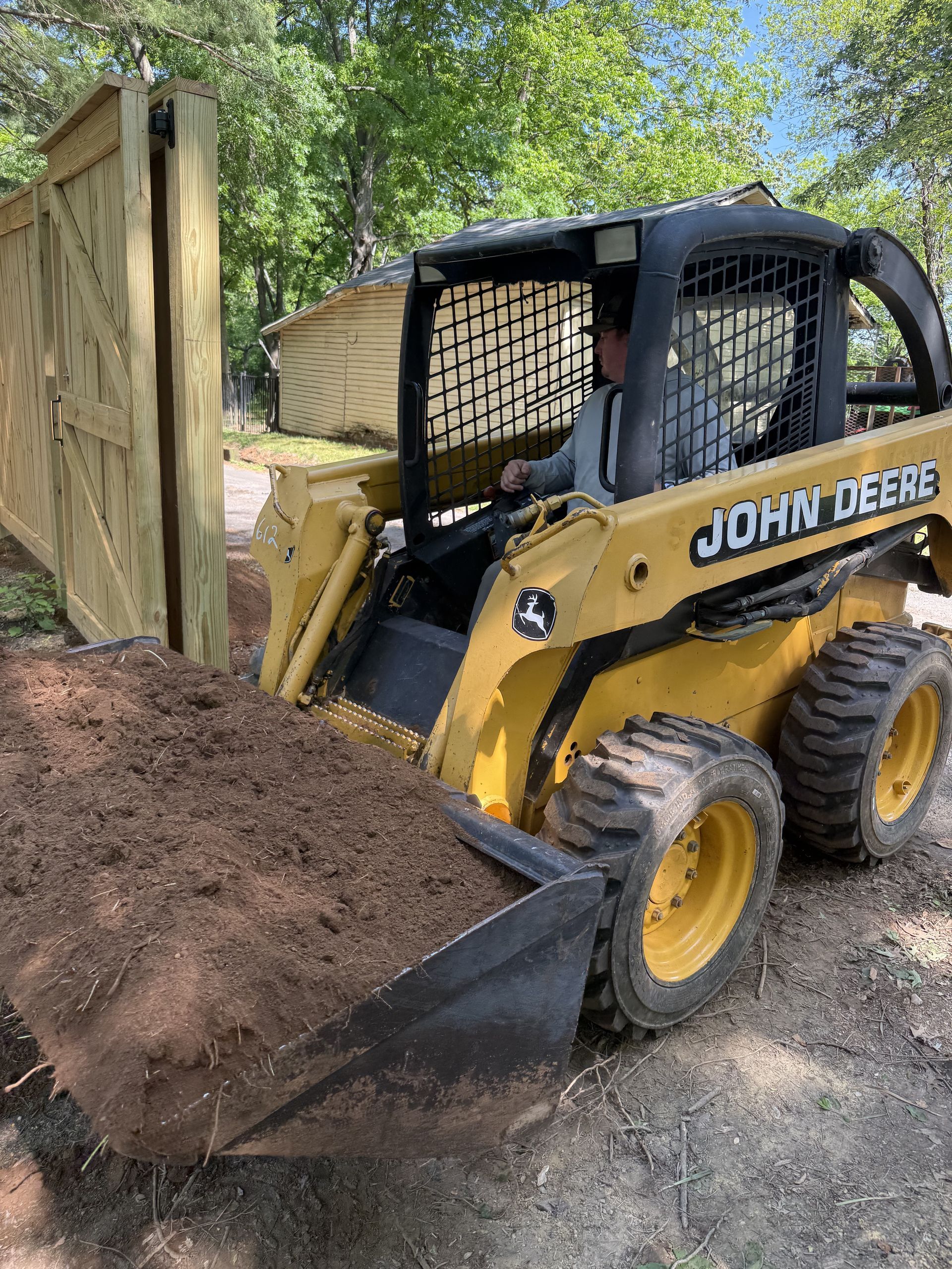 Yellow John Deere skid steer pushing dirt beside a wooden fence and shed in a wooded yard