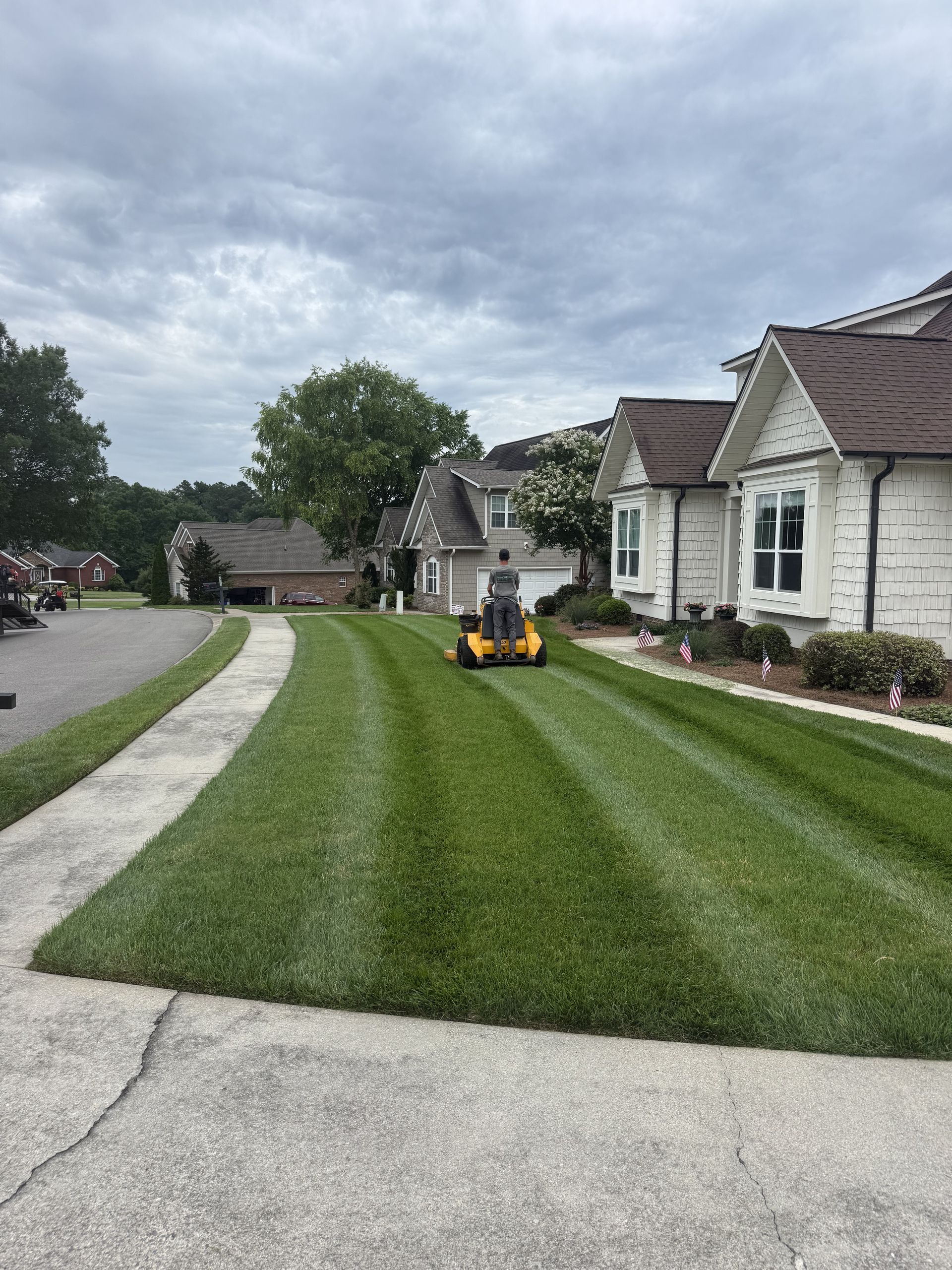 Suburban street with a striped green lawn and a yellow mower in front of houses under cloudy skies