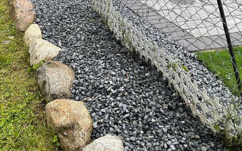 Rocks line a gravel path bordering a wire-enclosed rock wall and green grass.