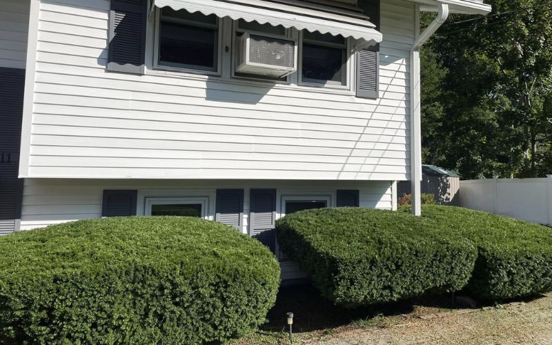 White house with white siding, dark shutters, air conditioner, and green hedges.