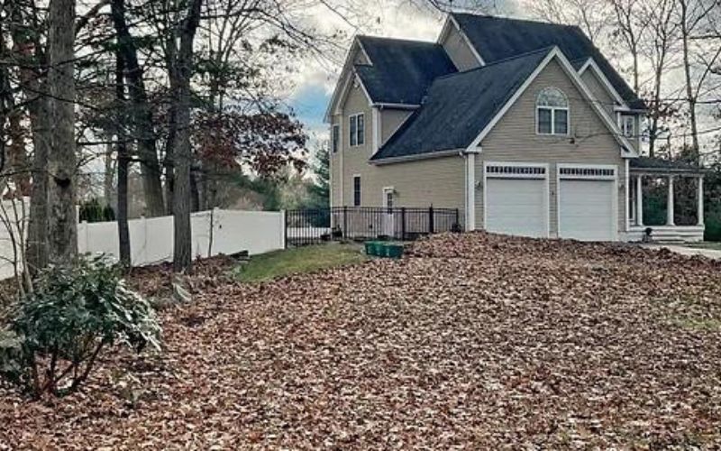 House with garage, yard covered in fallen leaves, surrounded by trees and white fence.
