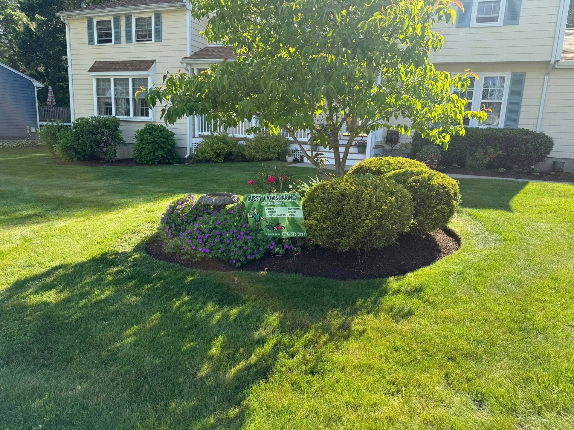 A landscaped front yard with a flower bed and a house in the background. Green grass, trees, and shrubs.