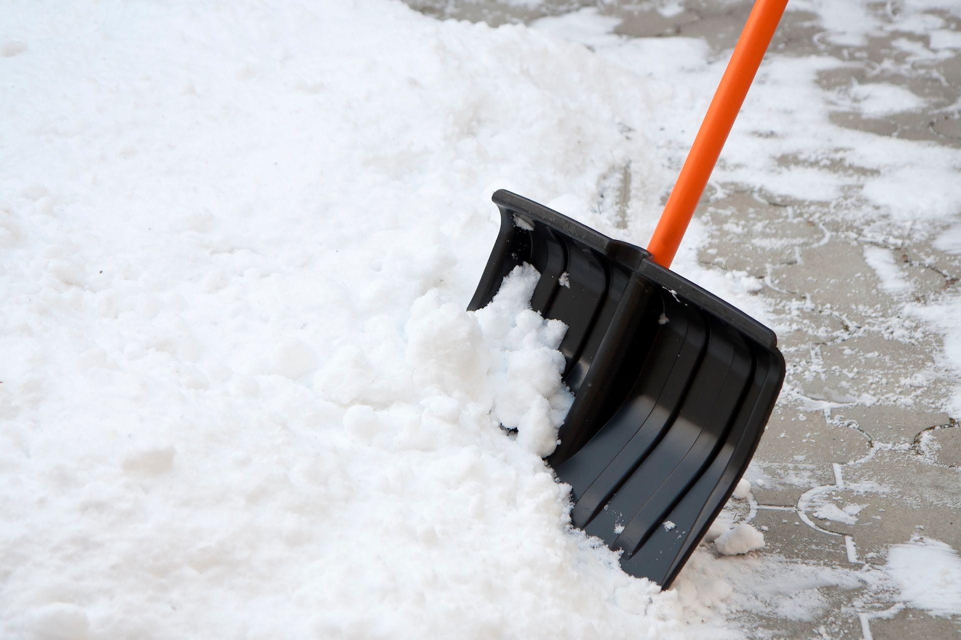 Three people shoveling snow from a sidewalk on a street lined with shops and parked cars; overcast day.
