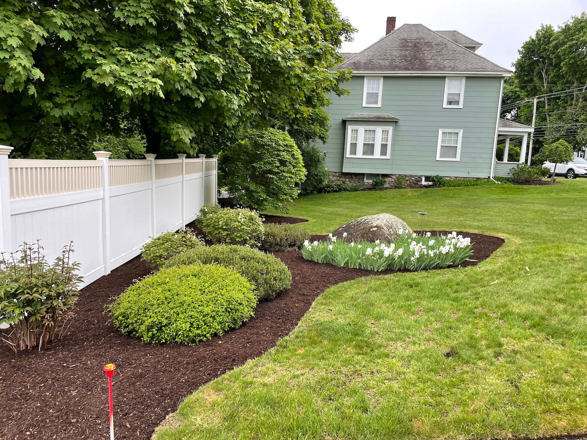 A green house with a white fence and a garden with mulch and flowers in the yard.