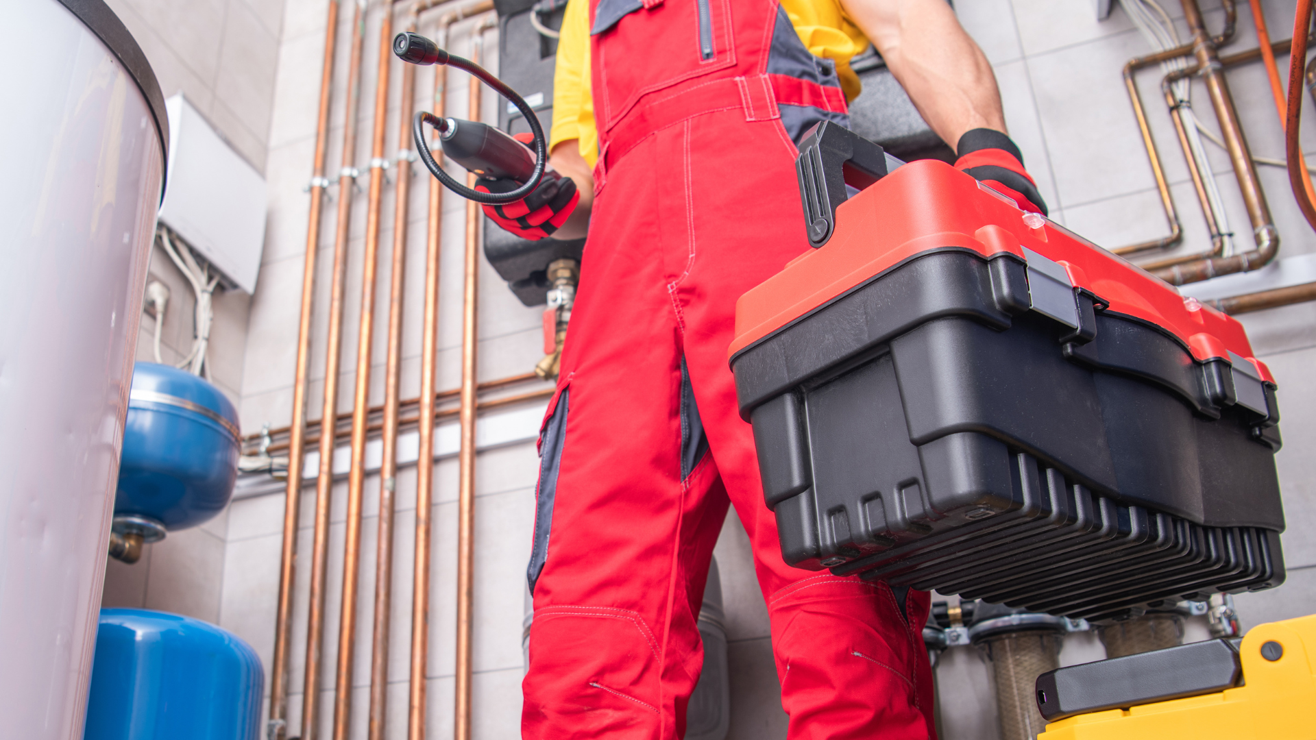 A plumber is holding a toolbox in a room with pipes.