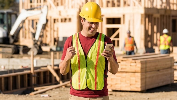 A construction worker is looking at his cell phone at a construction site.