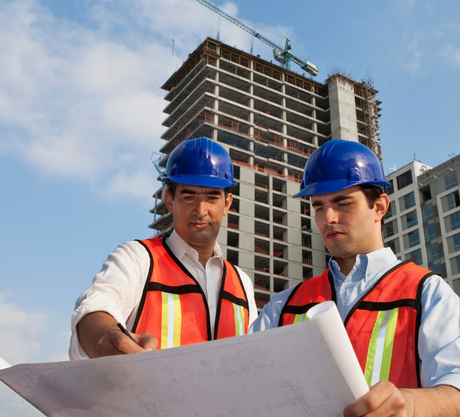 Two construction workers are looking at a blueprint in front of a building under construction