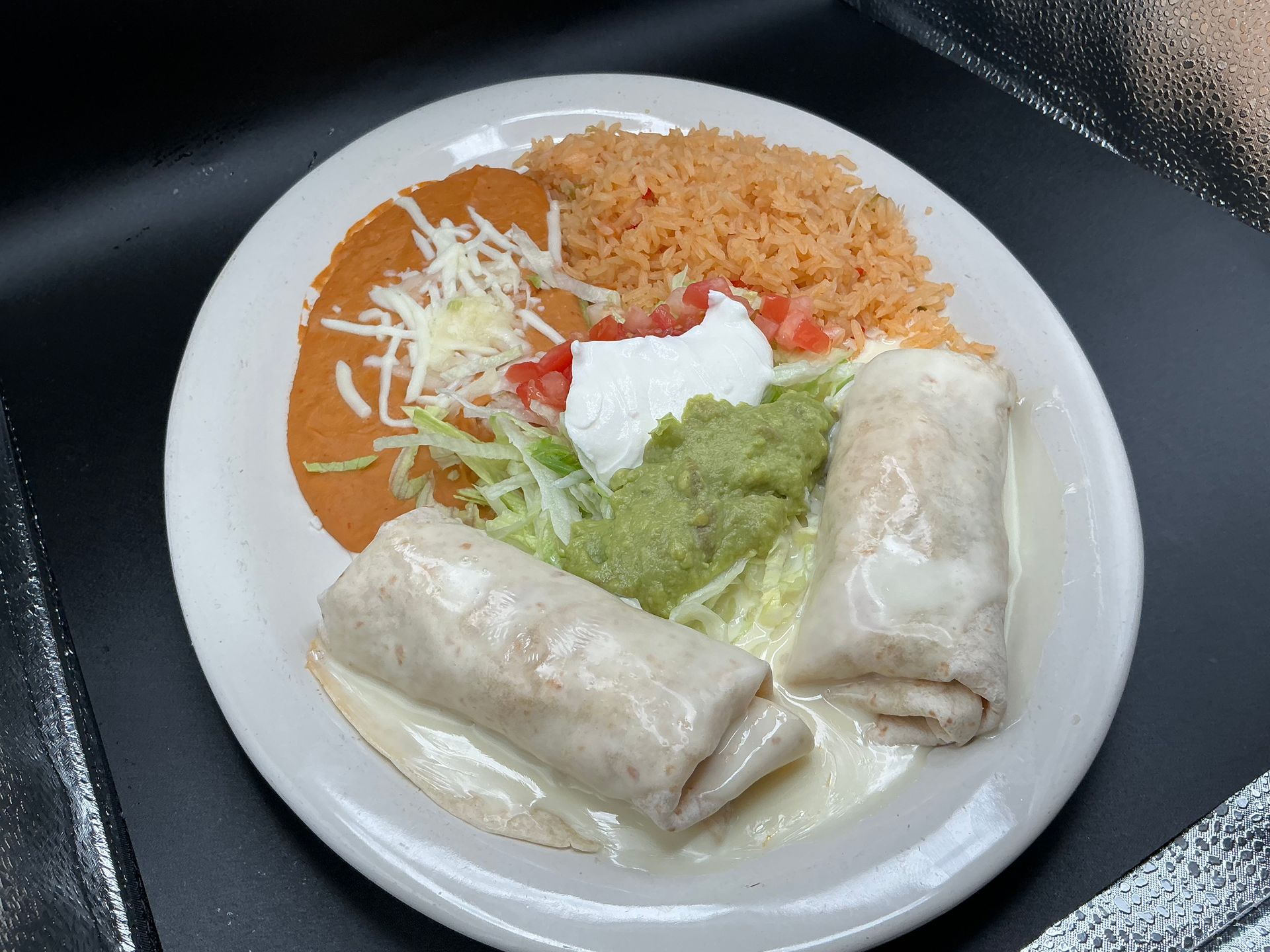 Plate of Mexican food: two burritos, refried beans, rice, guacamole, sour cream, lettuce, and tomatoes.