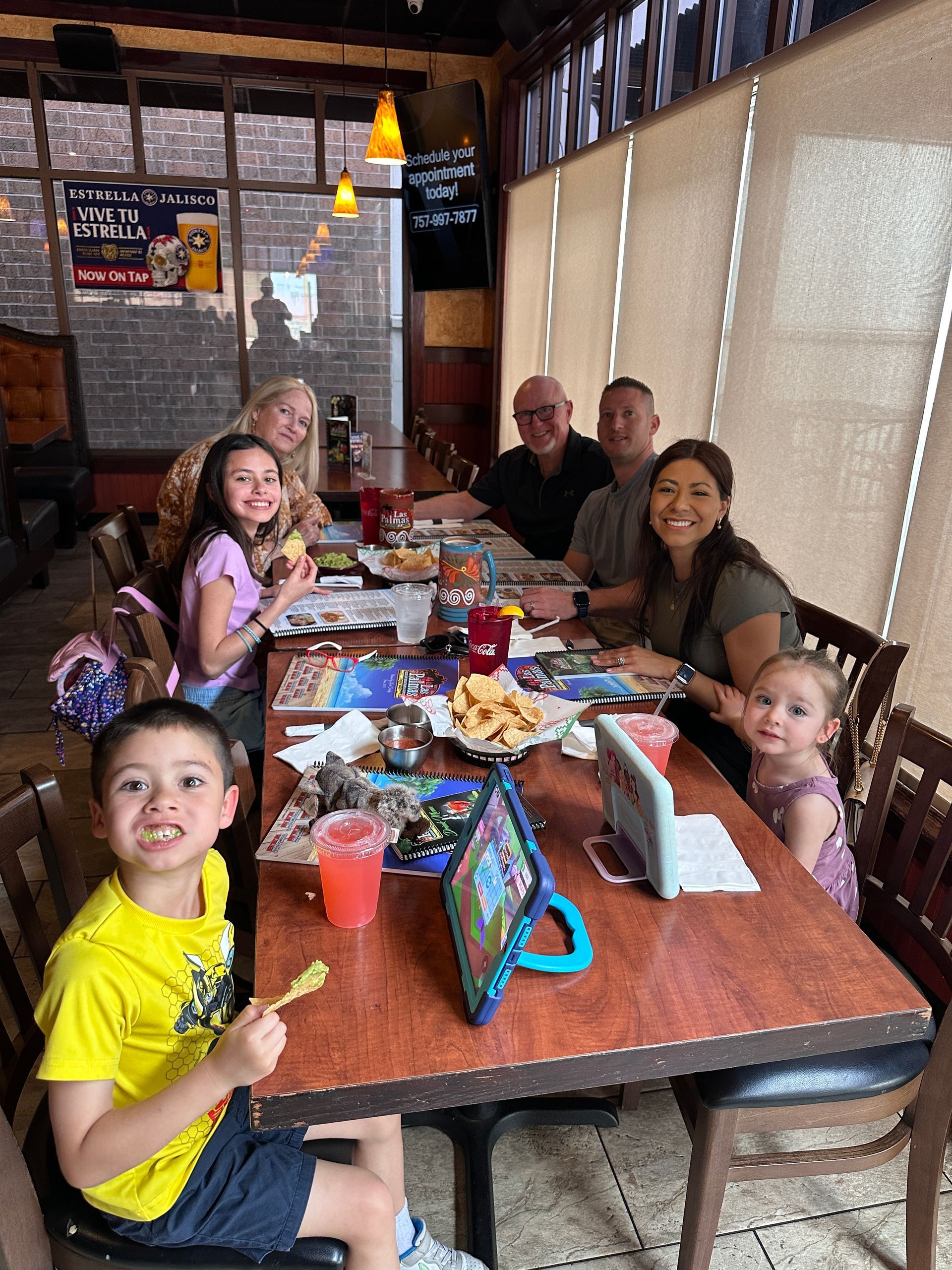 Family seated at a restaurant table, eating and smiling. Wooden table, food, and drinks visible.