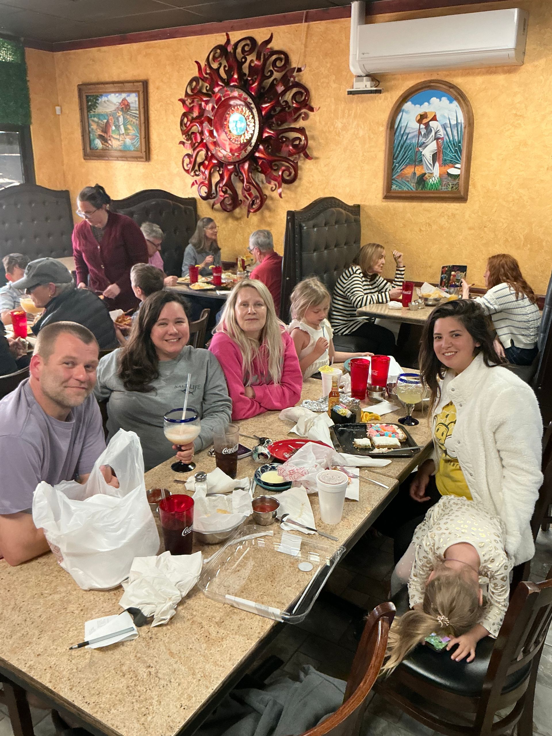 People at a restaurant table, dining with drinks and food. Decorative wall art, warm lighting.
