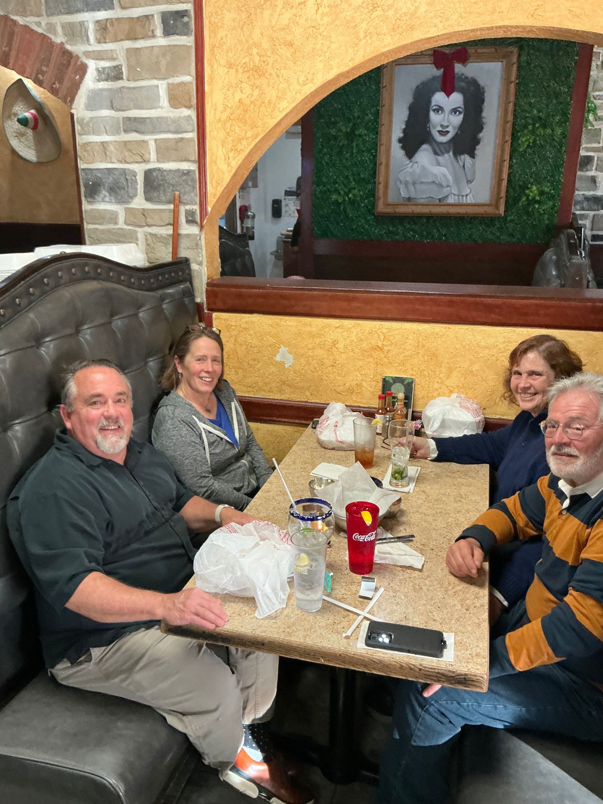 Four people sit at a restaurant table. One man smiles, others are talking. A portrait hangs in the background.