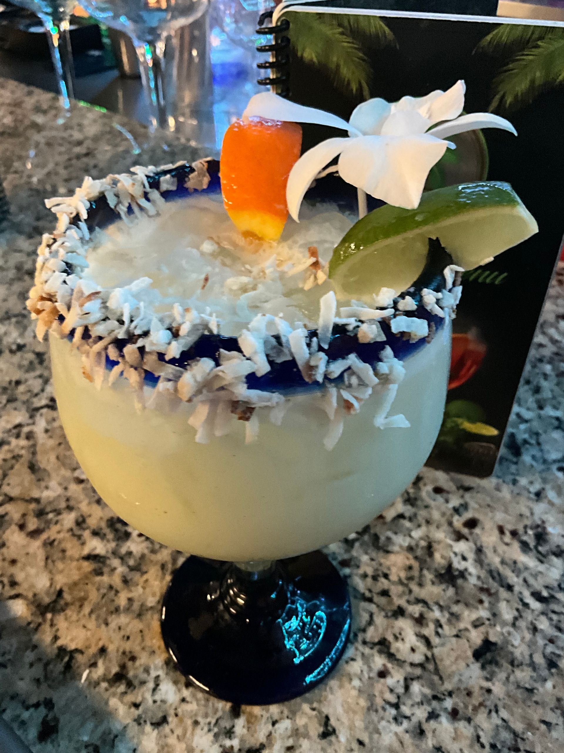 Coconut margarita in a blue-rimmed glass, garnished with lime, orchid and candy corn, sitting on a countertop.