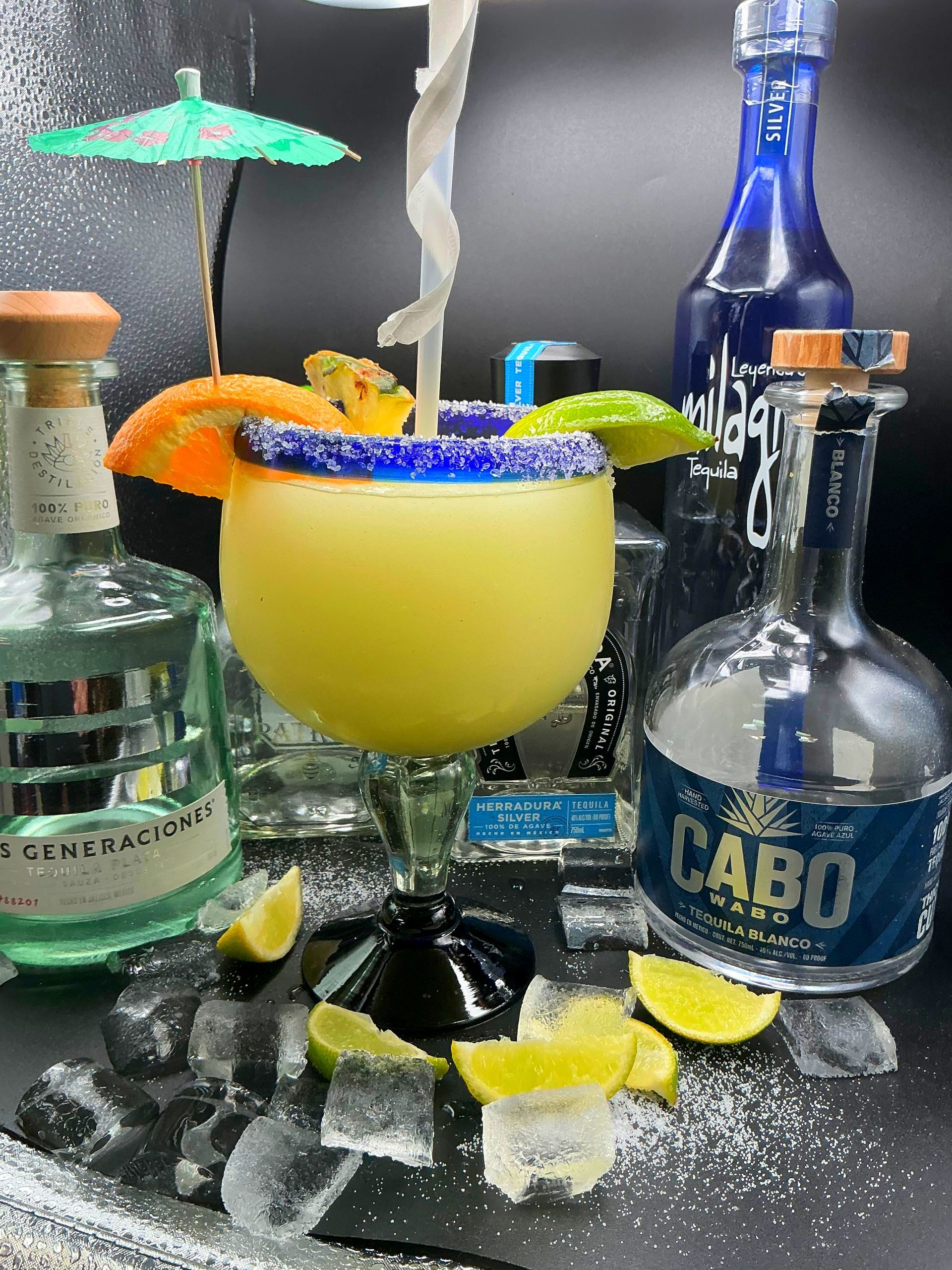 Margarita cocktail in a glass rimmed with blue salt, surrounded by tequila bottles, lime wedges, and ice.