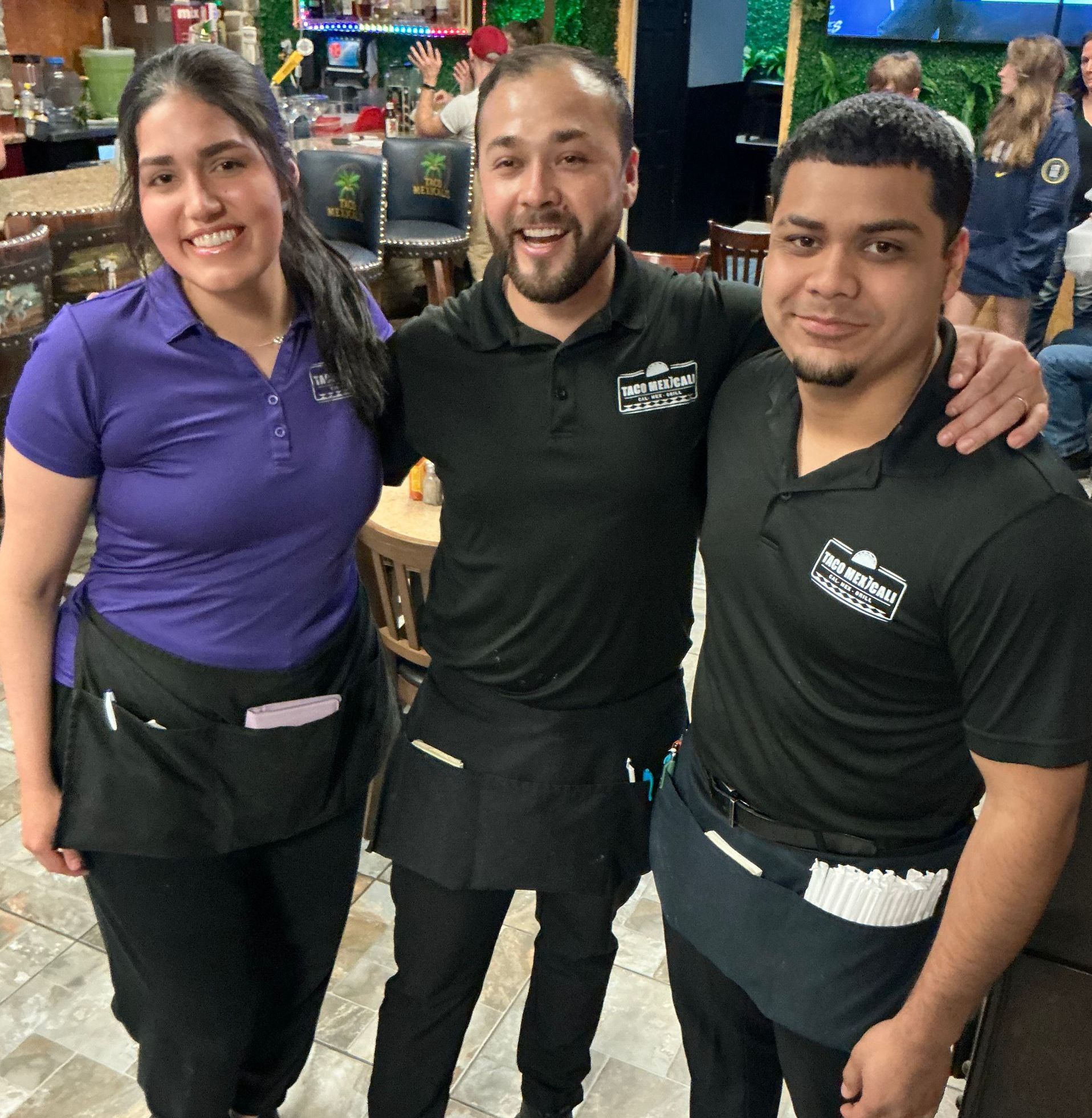 Three restaurant workers smiling, posing indoors. One woman in purple, two men in black, all with aprons.