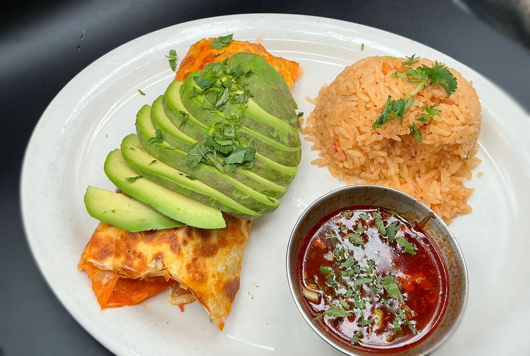 Plate with fried food, avocado slices, rice, and a small bowl of sauce.
