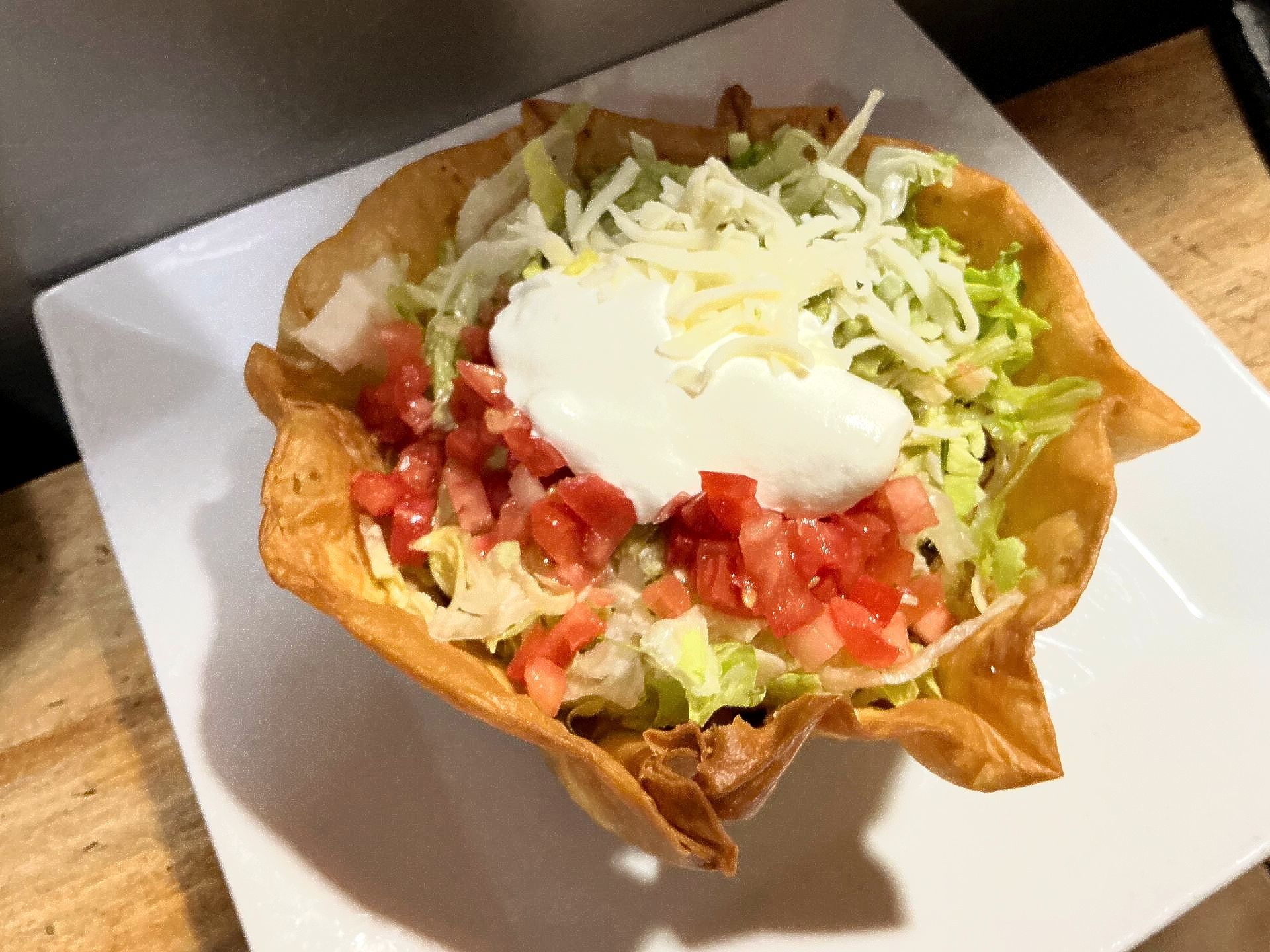 Taco salad in a crispy shell, filled with lettuce, tomatoes, cheese, and a dollop of sour cream on a white plate.