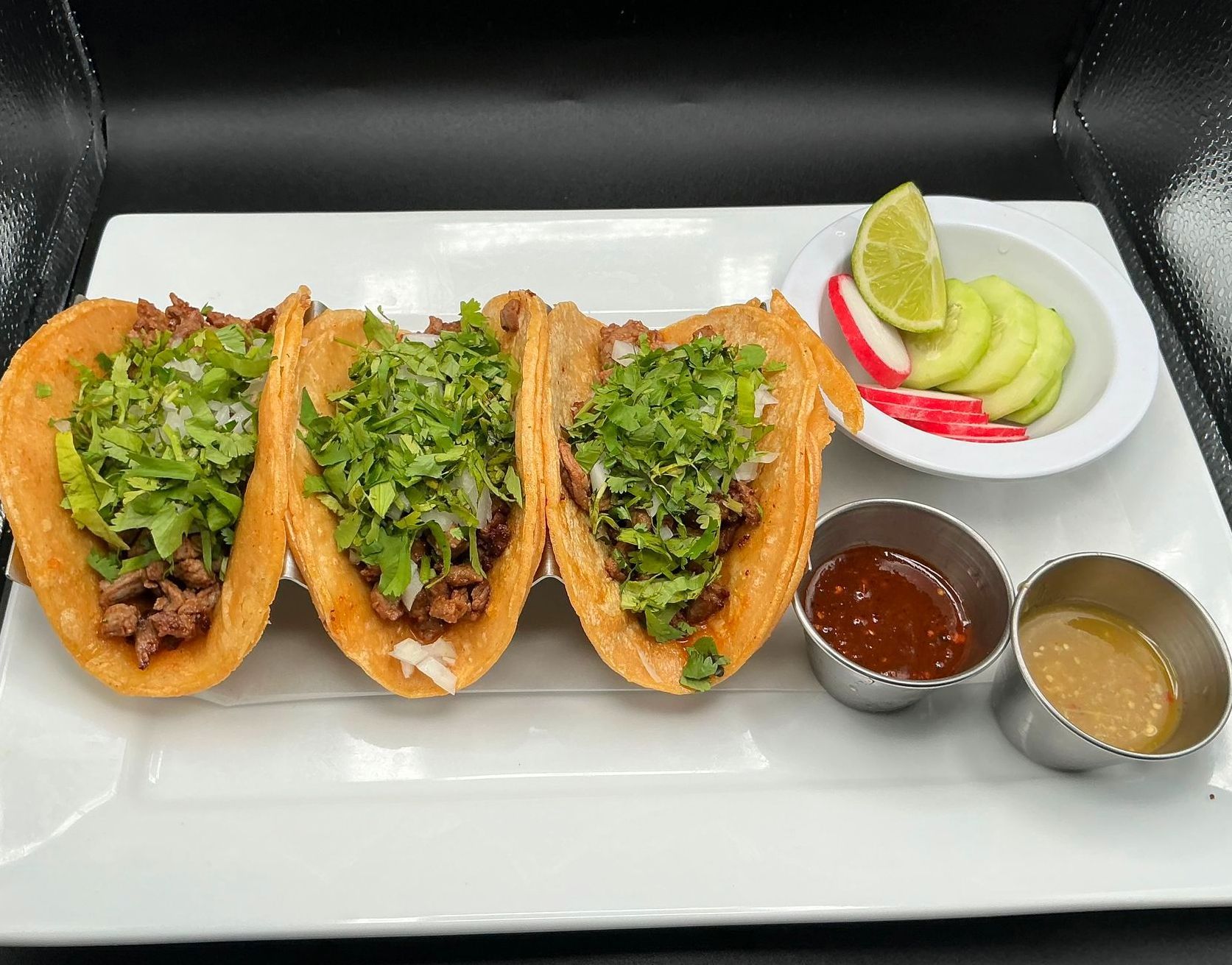 Three tacos on a white plate with salsa, lime, and cucumbers.