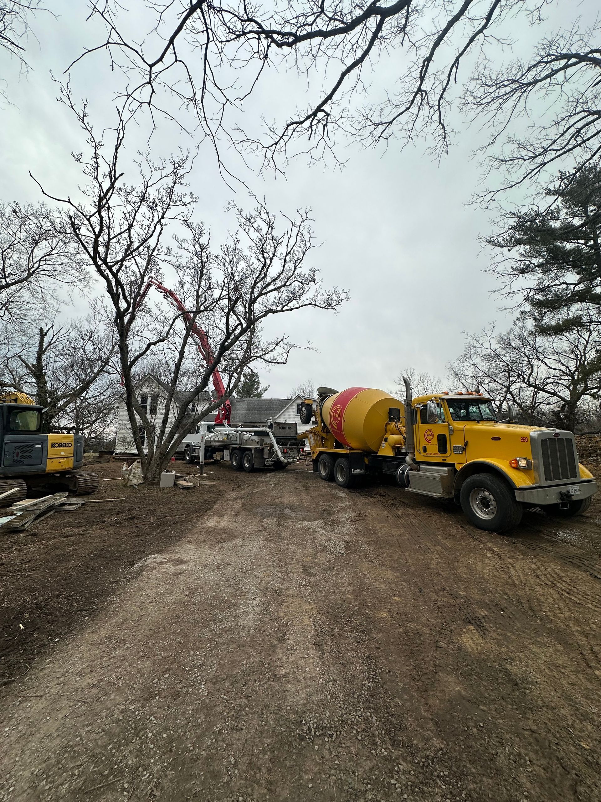 Yellow utility trucks and cement mixer on a muddy road lined with bare trees under a gray sky