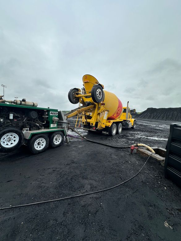 Yellow cement mixer overturned on a wet construction site beside a green truck under a gray sky