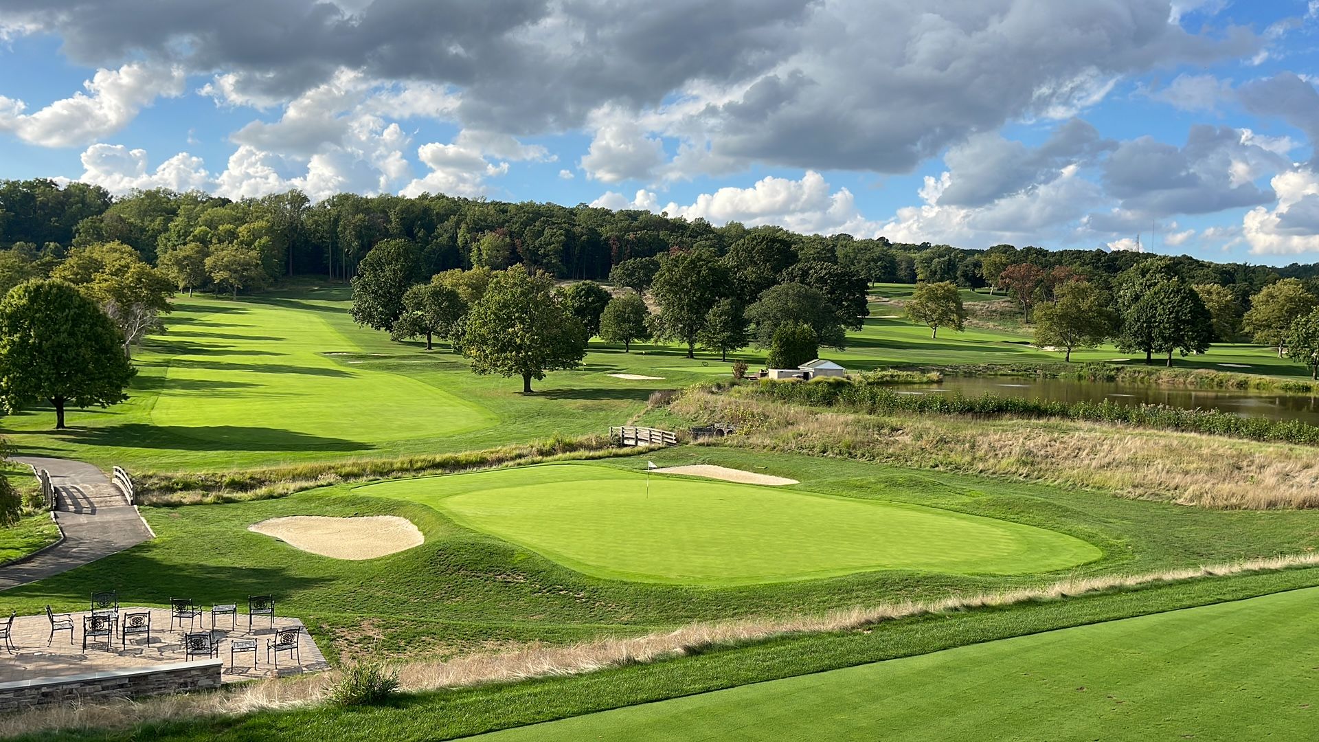 Green golf course with trees, water, and sand traps under a cloudy sky.