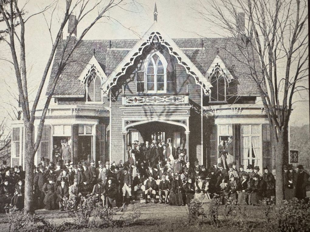 Large group of people pose in front of a house with elaborate Gothic Revival architecture; trees frame the scene.