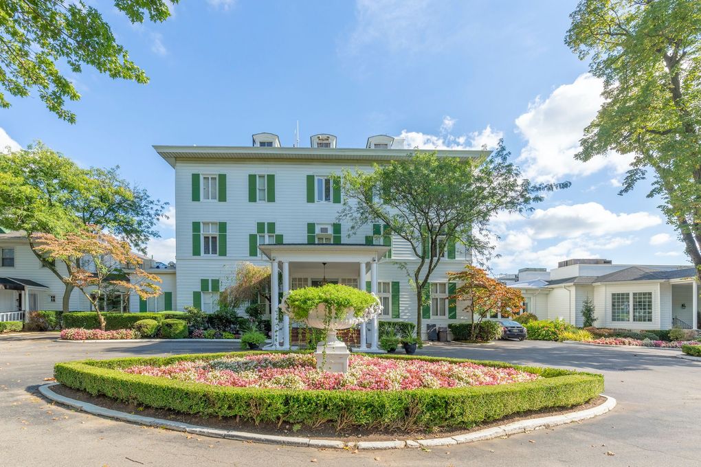 White building with green shutters, surrounded by trees and a flower bed. Sunny day.