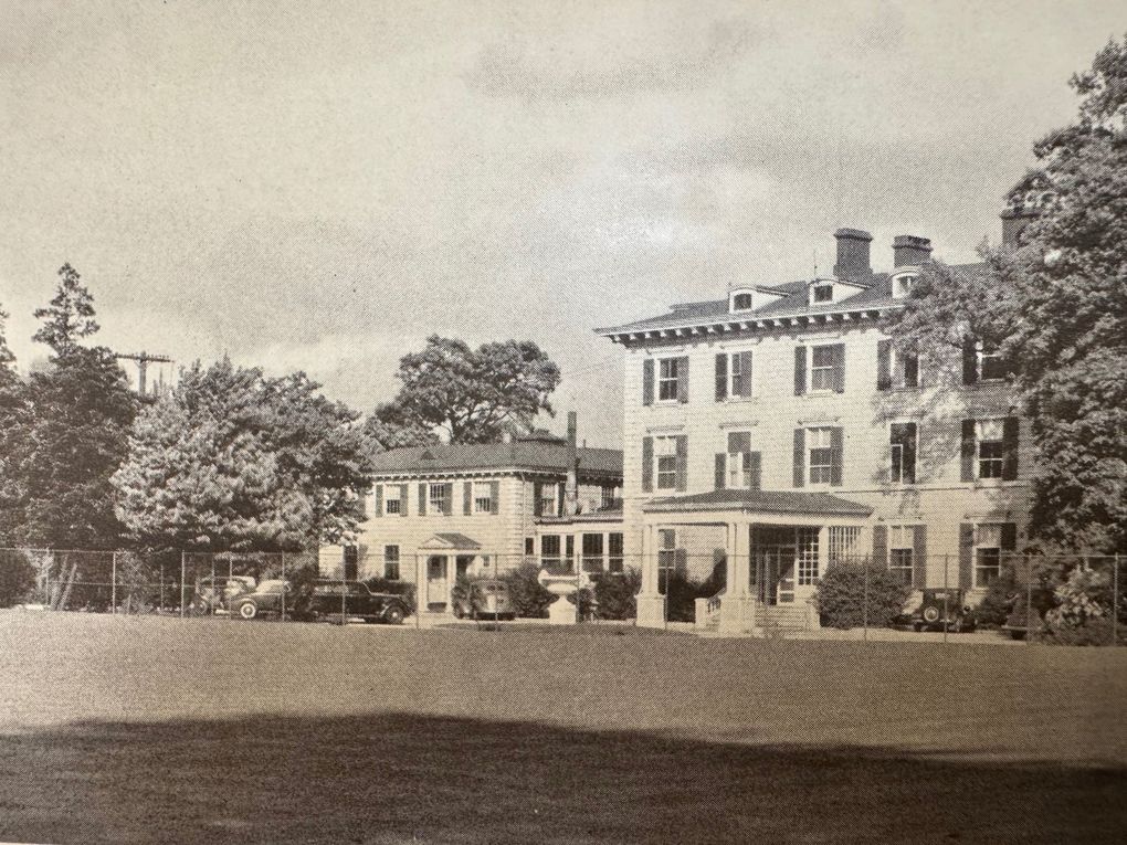Large, three-story house with porch and smaller connected building, set on a lawn with trees.