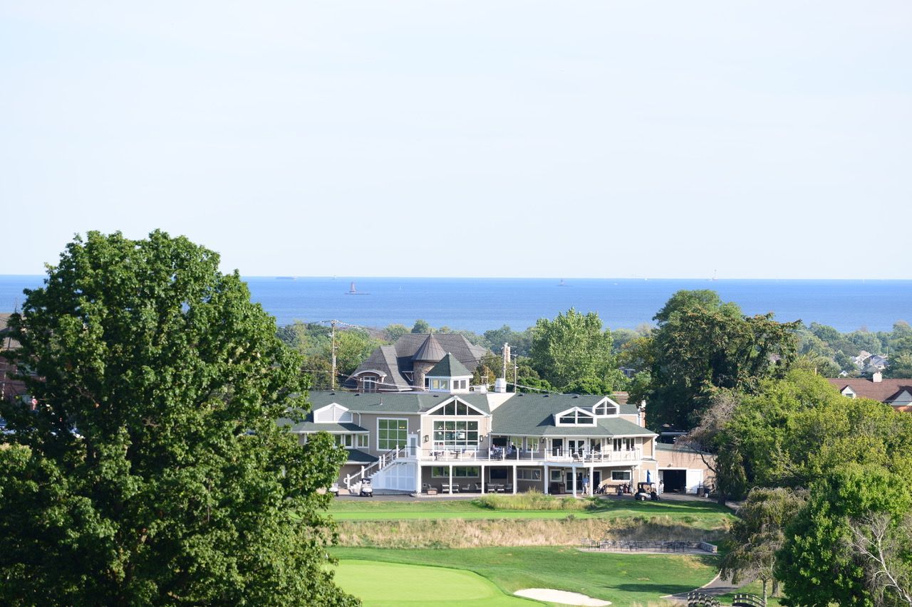 A large golf clubhouse with a long porch overlooking a golf course and a distant body of water.